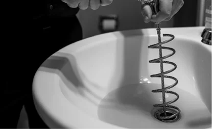 Person using a plumbing snake tool to unclog a bathroom sink drain. The coil of the snake is being inserted into the drain opening. The image is in black and white.