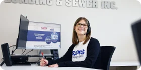 A woman wearing glasses and a headset smiles while sitting at a desk with a computer monitor. She holds a pen and wears a Capps Plumbing & Sewer shirt; part of the company name is visible on the wall behind her.
